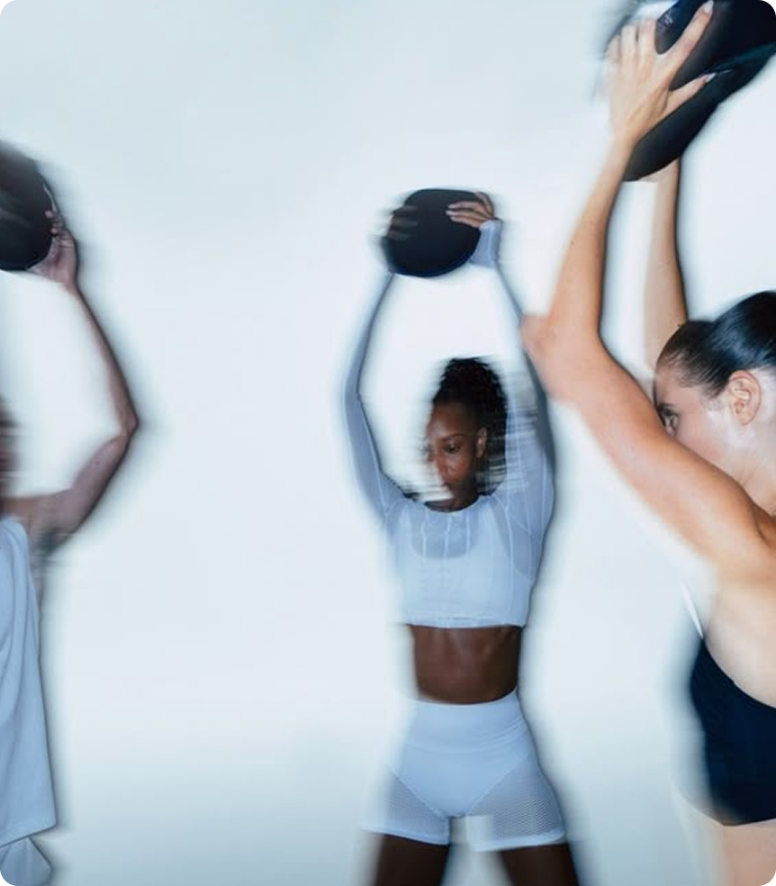 Zwei Frauen in Sportbekleidung die sich nebeneinander Bewegen und Langhanteln nutzen. Das bIld nutzt viel Bewegungsunschärfe für Dynamik und Stärke.
