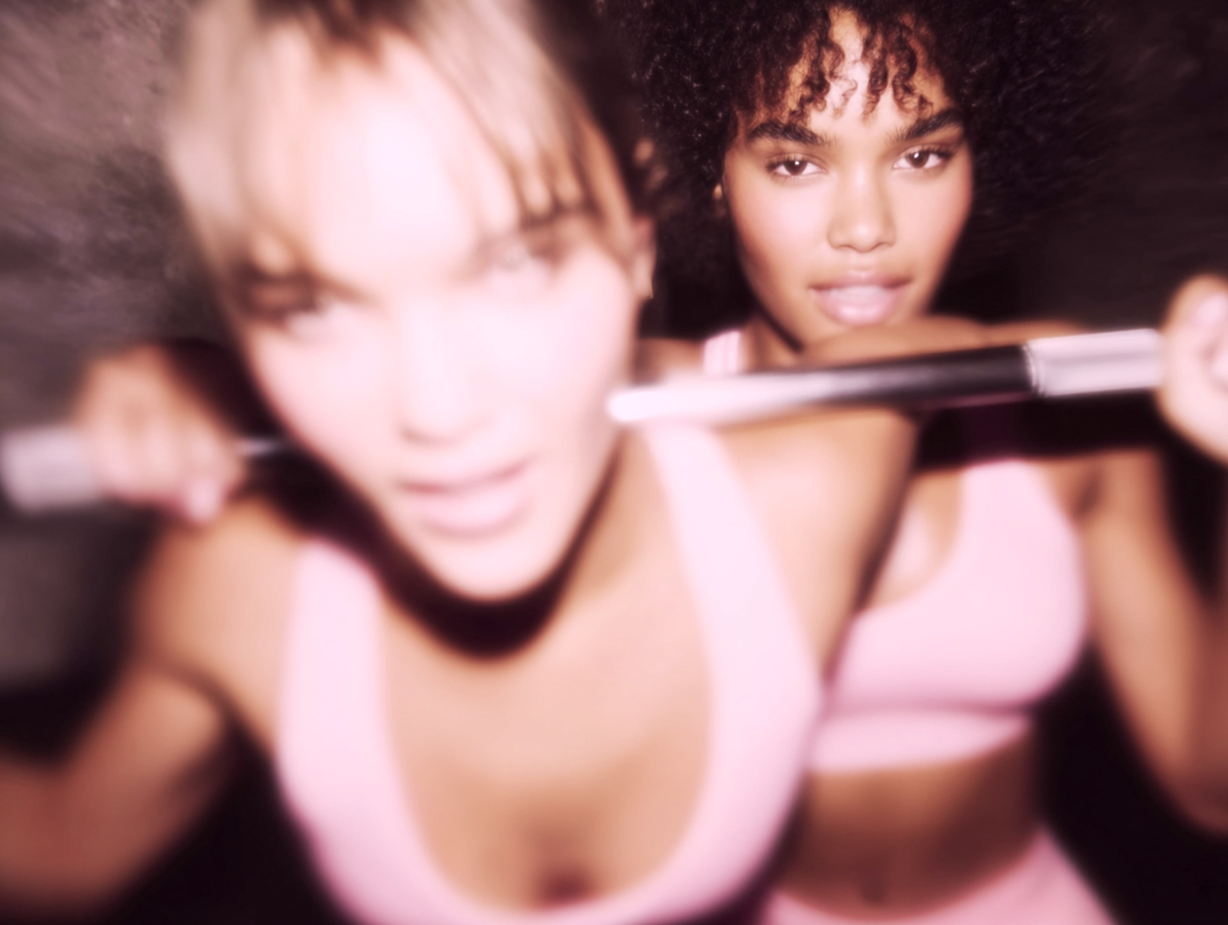 Eine Nahaufnahme der Gesichter von zwei Frauen in rosa Sportbekleidung die in einem Fitnessstudio Kraft trainieren.